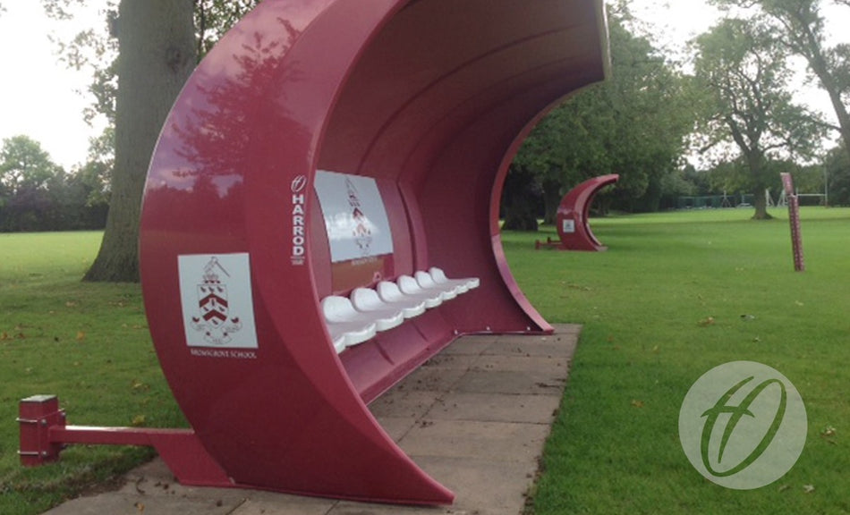 Harrod Sport Fibretech "Pro" Shelter Anchors in maroon, with white seats and support posts, are positioned on a grassy sports field. The semi-enclosed benches are branded with Harrod logos. Trees and another shelter appear in the background.