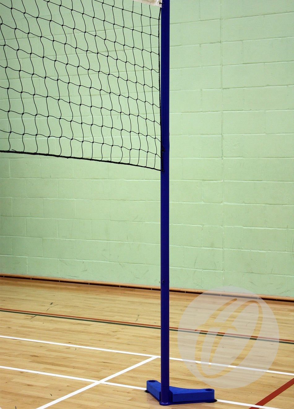 A Harrod Sport Floor Fixed Schools Volleyball Post, featuring a blue powder-coated finish, is installed on a wooden indoor court with a light green brick wall in the background. The attached volleyball net is partially visible.