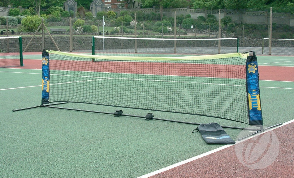 The Harrod Sport Foldaway Mini Tennis net, featuring a sturdy PVC frame, is set up on outdoor green and red courts; a carry bag sits close by, with several other tennis courts visible in the background.