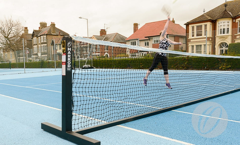 A person on a blue outdoor tennis court jumps near the regulation net, supported by Harrod Sport Freestanding Tennis Posts (Black), to hit a tennis ball, with houses and trees in the background under a cloudy sky.