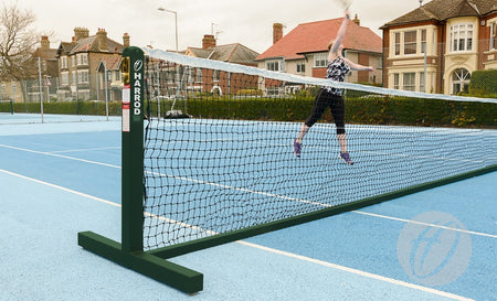 Wearing athletic clothing, a player jumps to hit a tennis ball over the net on an outdoor blue court equipped with Harrod Sport Freestanding Tennis Posts (Green), with residential houses visible in the background.