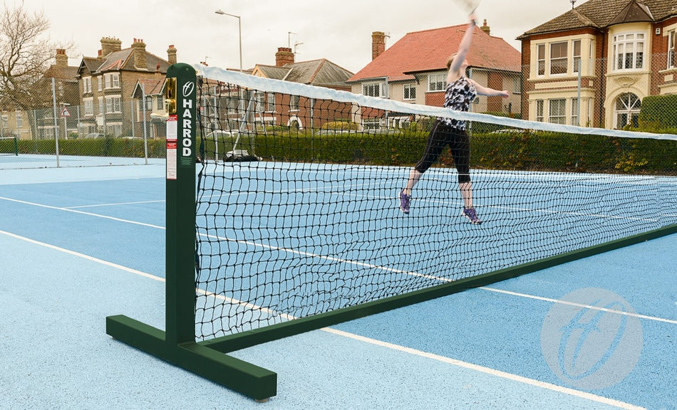 Wearing athletic clothing, a player jumps to hit a tennis ball over the net on an outdoor blue court equipped with Harrod Sport Freestanding Tennis Posts (Green), with residential houses visible in the background.