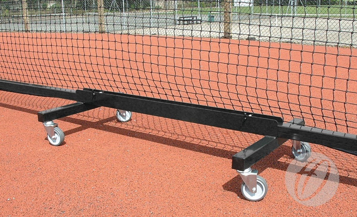 A close-up of Harrod Sport Freestanding Tennis Trolleys - Black, each with four caster wheels, on a red outdoor court. The trolleys resemble wheeled frames; more fencing and an out-of-focus outdoor area are visible in the background.