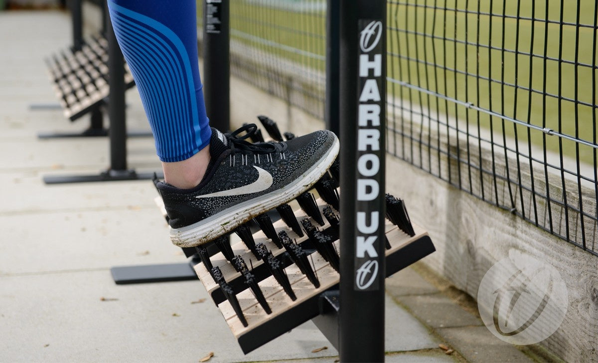 Wearing blue leggings, someone stretches their calf on a metal Harrod Sport Socketed Angle Boot Wiper beside beech timber boot cleaning brushes, near a fenced sports field, while wearing black and white Nike running shoes.
