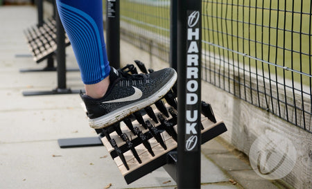 Wearing blue leggings, someone stretches their calf on a metal Harrod Sport Socketed Angle Boot Wiper beside beech timber boot cleaning brushes, near a fenced sports field, while wearing black and white Nike running shoes.