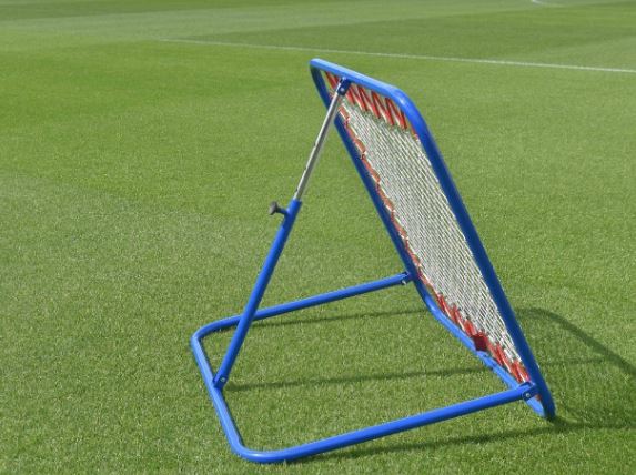 A Samba Sports Pro Rebounder with a blue frame and red accents is positioned on a green sports field, perfect for ball training drills and first touch practice.
