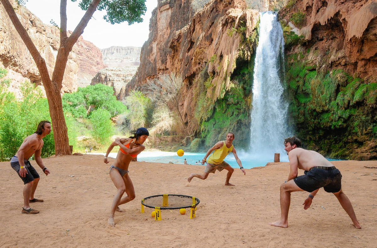 Four people in swimwear play Kankakee Spikeball Standard Set, an exciting outdoor game, on sand near a waterfall and rocky cliffs, with lush trees and blue water in the background.