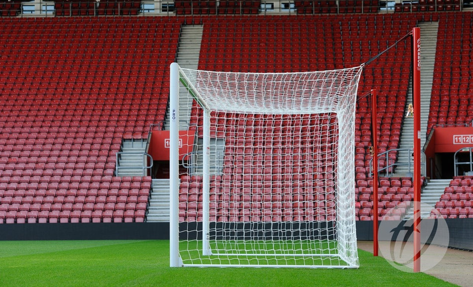 A Harrod Sport Stadium Pro Bottom Hinged Net Support with netting and an aluminium bar stands on a green field before empty red stadium seats in a well-maintained, match-ready arena with no people visible.