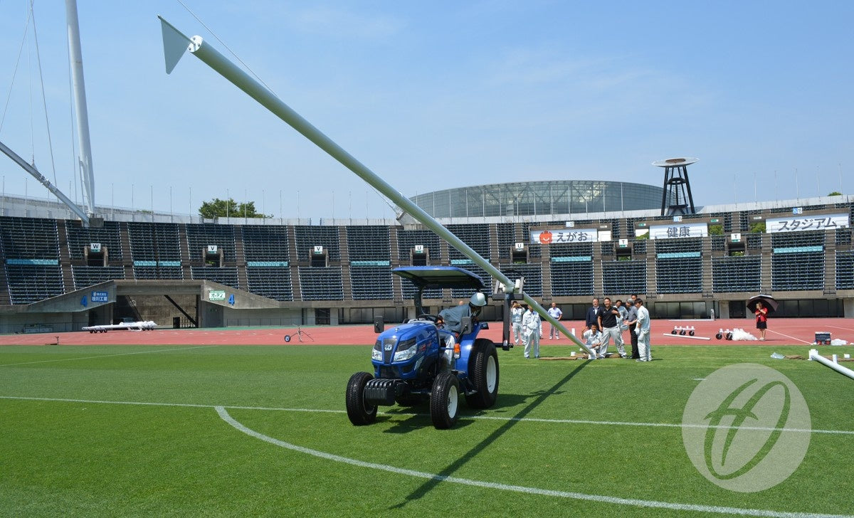 A Harrod Sport Hinged Rugby Post Lifter, attached to a blue tractor on a stadium field, supports an upright pole as several people gather close by; empty seats and the stadium structure appear in the background.