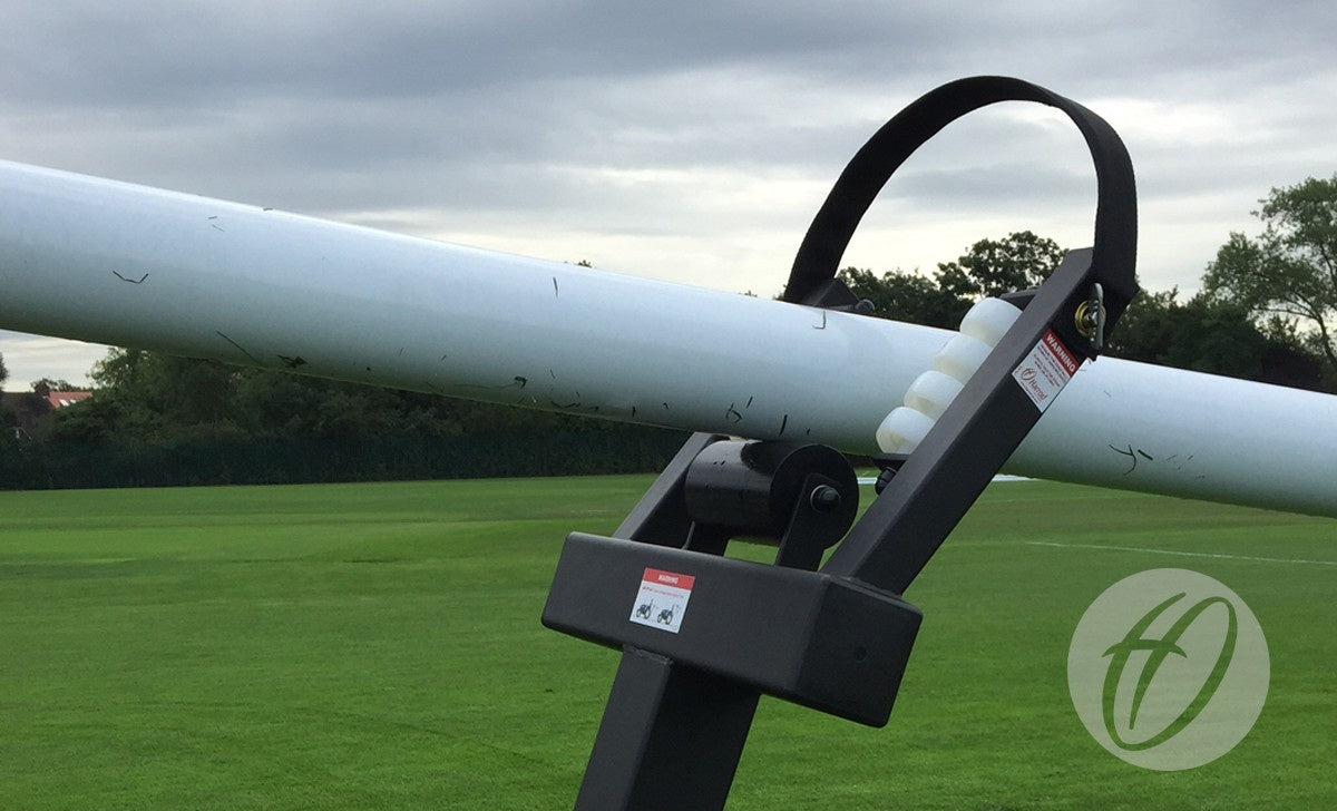 A close-up of the Harrod Sport Hinged Rugby Post Lifter, featuring a white cylindrical bar and black handle, on a grassy field with trees and a cloudy sky in the background.