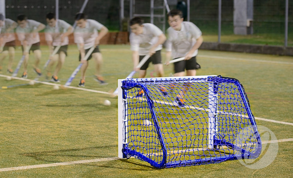 A person quickly swings a hockey stick, striking a ball toward a small blue and white Harrod UK Hockey Target Goal by Harrod Sport on an outdoor field, with motion blur highlighting the action.