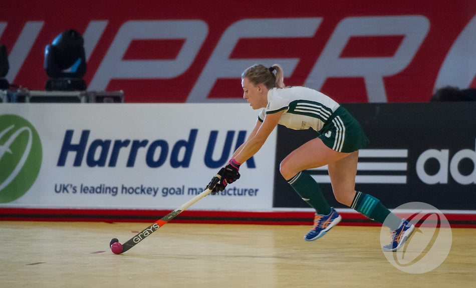 A female field hockey player in green shorts and a white jersey lunges forward with her stick to hit a pink ball on an indoor court lined with Harrod Sport Aluminium Indoor Hockey Pitch Sideboards.