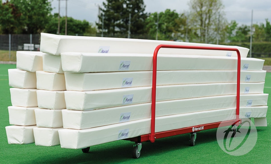 A red Harrod Sport Hockey Rebound Storage Trolley stacked with white foam pads sits on a green outdoor surface, with trees and a fence in the background—ideal for transporting Hockey Pitch Divider Pads.