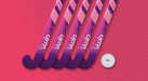 Five Uwin TS-X Hockey Sticks in pink and purple are arranged diagonally on a textured pink surface, with a white field hockey ball placed nearby.