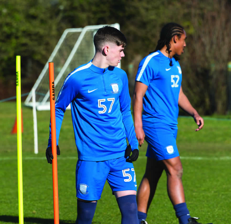 Two soccer players in blue kits (numbers 57 and 52) stand on grass near Samba Sports Samba Junior Boundary Poles 1.5m (set of 12), with a goalpost and trees visible in the background.