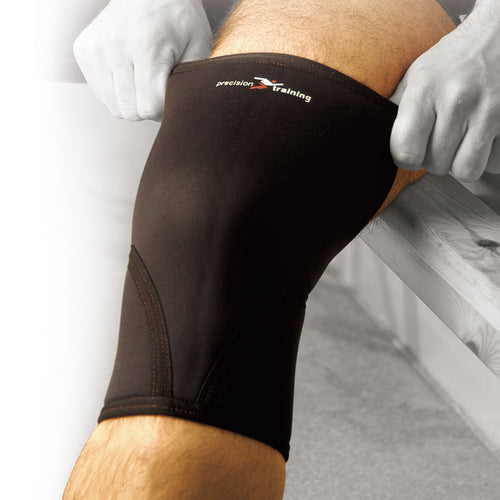 A person pulls the Precision Training Precision Neoprene Knee Support, in black, onto their knee while sitting on a bench for added arthritis relief.