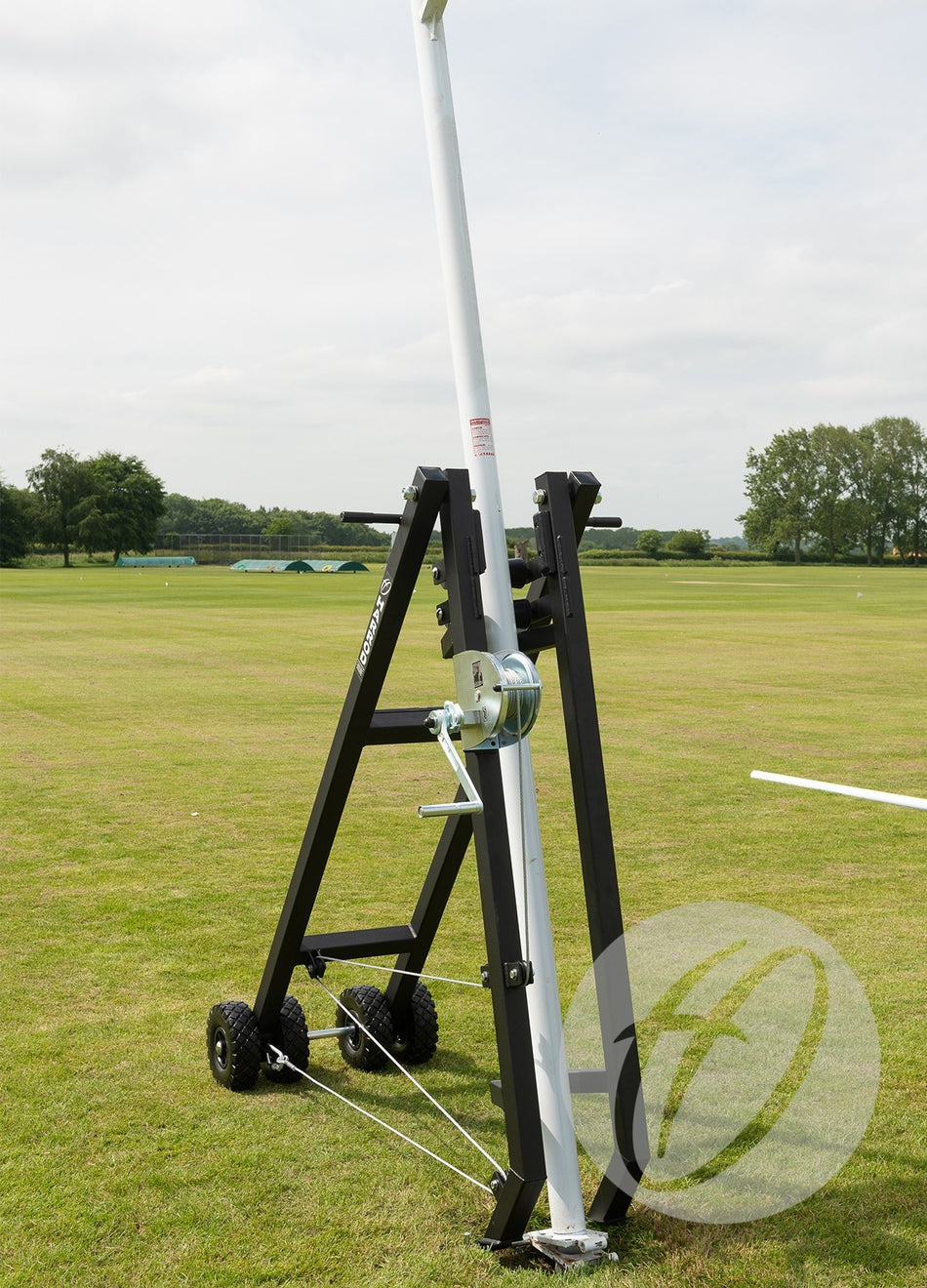 The Harrod Sport Winched Rugby Post Lifter, a portable system with wheels and a black frame, holds an aluminium rugby post upright on a sports field. Trees and a cloudy sky are visible in the background.