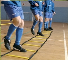 Four people in blue sports uniforms and black shoes practice agility drills using the Precision Training Foot Speed Ladder Indoor Weighted on a gym floor to enhance their foot speed.