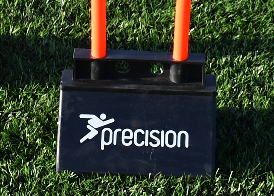 A close-up of a Precision Rubber Mini Mannequin Base by Precision Training with two orange poles, set on green artificial grass. The durable base features the Precision Training logo and name clearly visible.