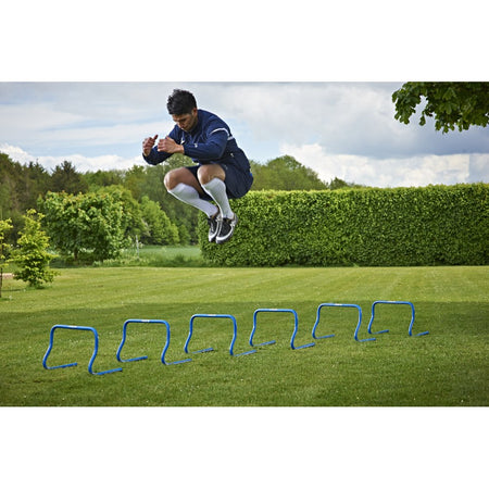 A man in athletic gear leaps over the Mitre Training Hurdle Set 12" by Mitre on a grassy lawn, demonstrating coordination drills surrounded by green trees and bushes under a partly cloudy sky.