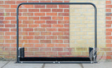 The Harrod Sport Fixed Multi Boot Wiper, featuring a polyester powder coated metal frame with black bristles and side brushes, is positioned on a paved area in front of a brick wall as a sturdy boot cleaning station.
