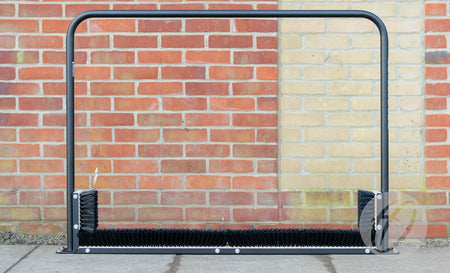The Harrod Sport Fixed Multi Boot Wiper, featuring a polyester powder coated metal frame with black bristles and side brushes, is positioned on a paved area in front of a brick wall as a sturdy boot cleaning station.