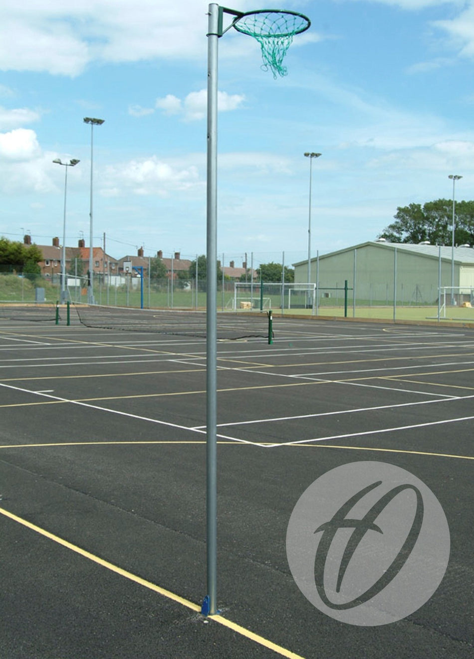 A Harrod Sport Regulation Netball Post - C/W Locking Sockets with a green net and steel uprights stands on an outdoor court with marked lines. Other courts, fencing, buildings, and tall floodlights appear under a partly cloudy sky.