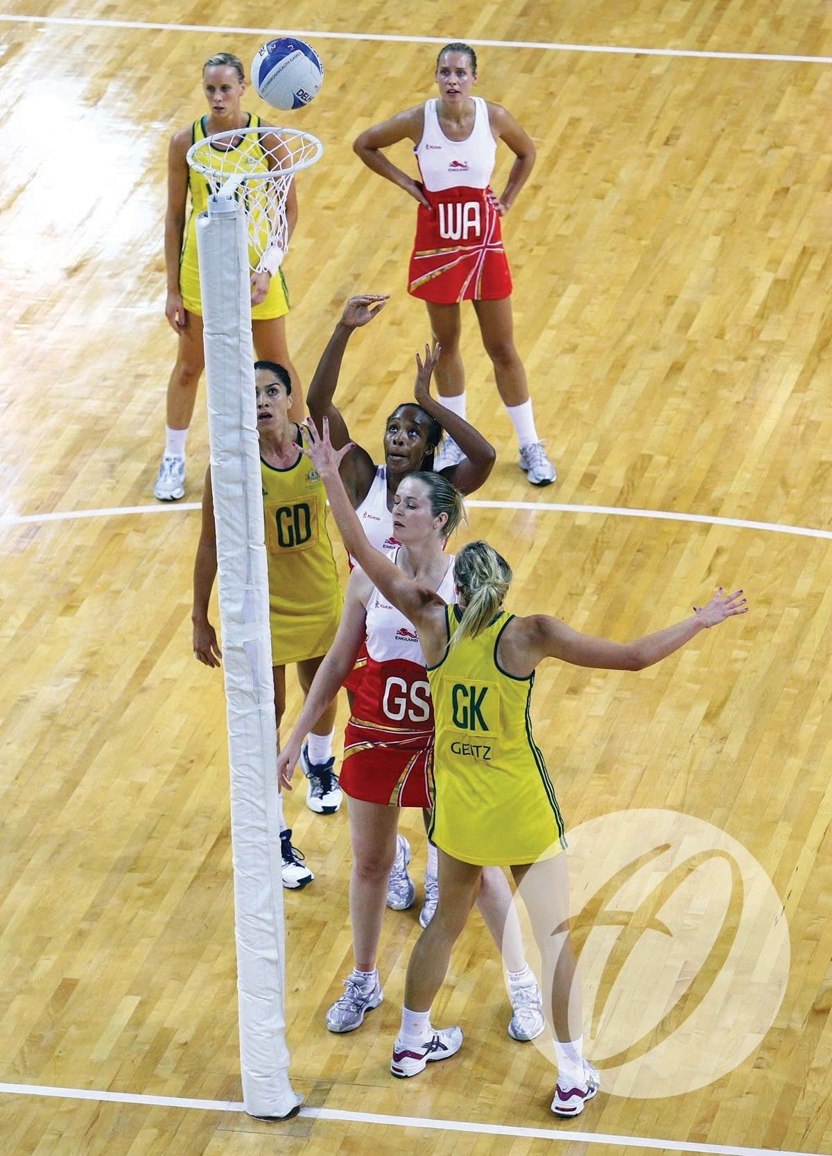 Players from two teams, one in yellow and one in red, compete around the goalpost fitted with Harrod Sport Competition Netball Protectors - Single Colour as the ball is about to score on a polished indoor court.