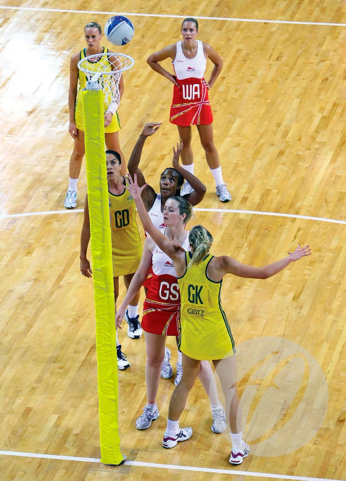 A netball game unfolds on an indoor wooden court, with players in yellow and red. The goalpost is fitted with Harrod Sport Competition Netball Protectors - Single Colour as one player attempts a shot.