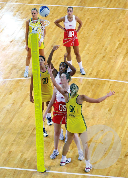 A netball game unfolds on an indoor wooden court, with players in yellow and red. The goalpost is fitted with Harrod Sport Competition Netball Protectors - Single Colour as one player attempts a shot.