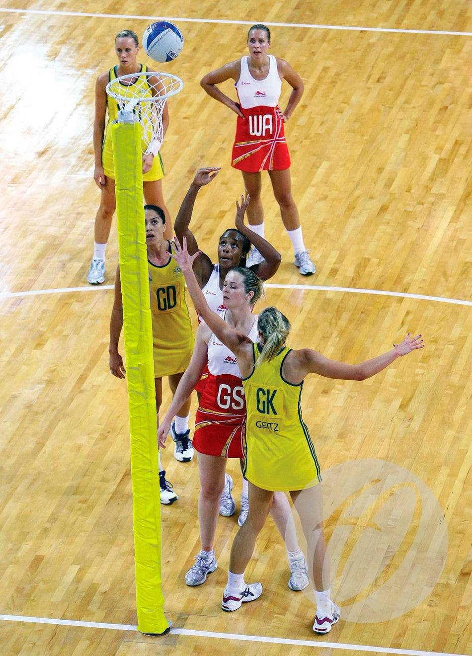 A netball game unfolds on an indoor wooden court, with players in yellow and red. The goalpost is fitted with Harrod Sport Competition Netball Protectors - Single Colour as one player attempts a shot.