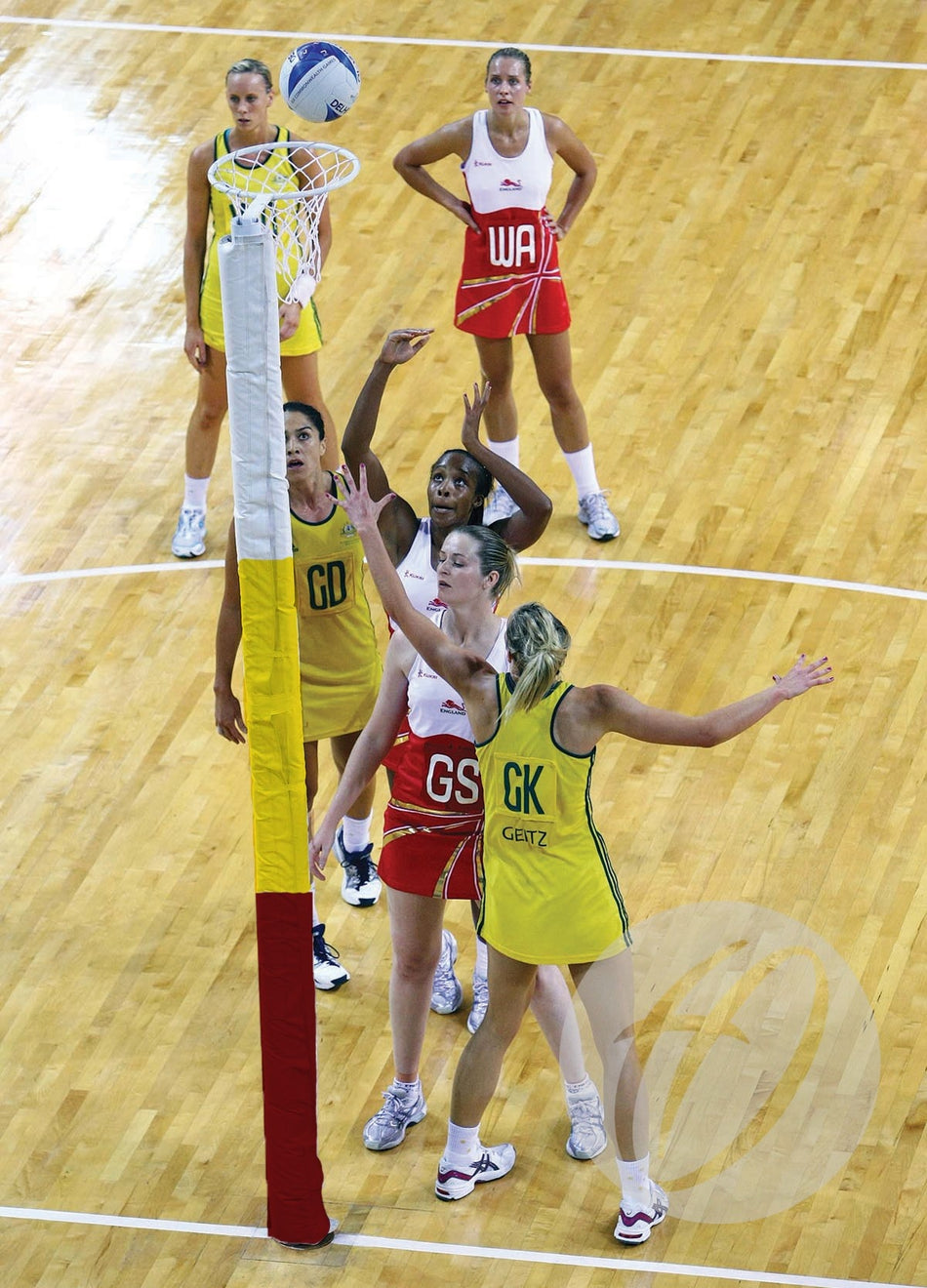 Four female netball players, two in yellow and two in red, stand under a netball hoop with Harrod Sport Competition Netball Protectors – Three Colour. A red player prepares to shoot as others watch or defend; two more players are visible in the background.