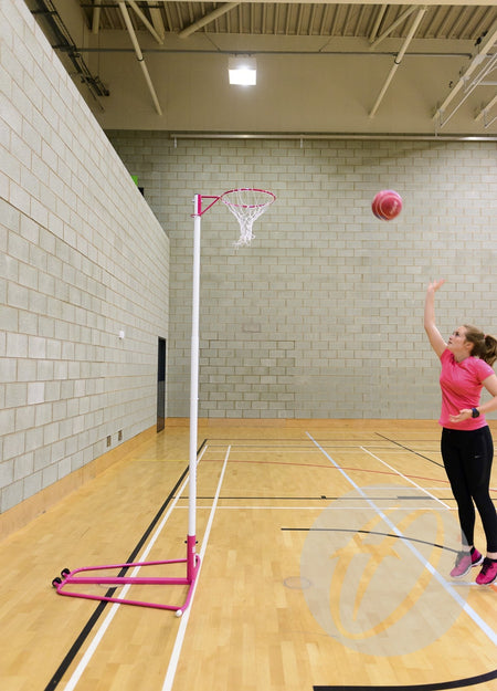 A woman in a pink shirt shoots a basketball toward the Harrod Sport Telescopic Netball Posts (Pair) in an indoor gym. The lightweight aluminium supports and net are also pink, complementing the light-colored brick walls.