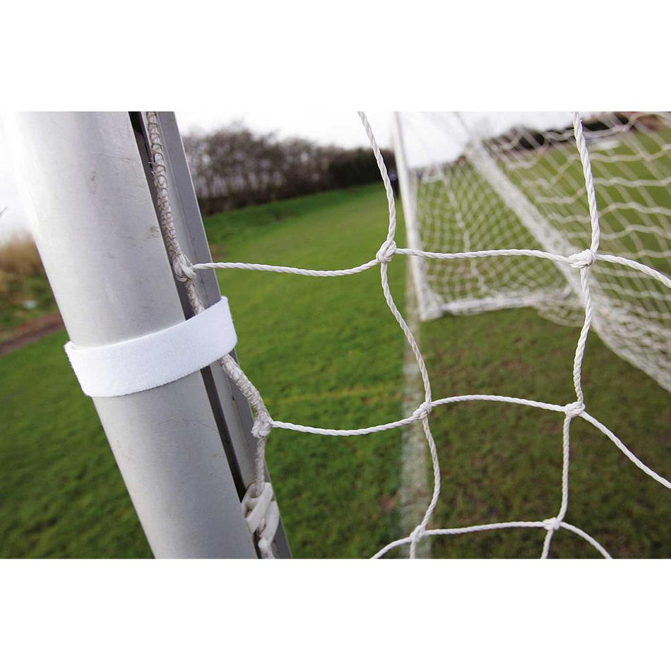 Close-up of a Precision Net Fabric Fastener from Precision Training securing a soccer net to a metal goalpost on a grassy outdoor field, with another goal and trees visible in the background. Sold as a roll of 24.