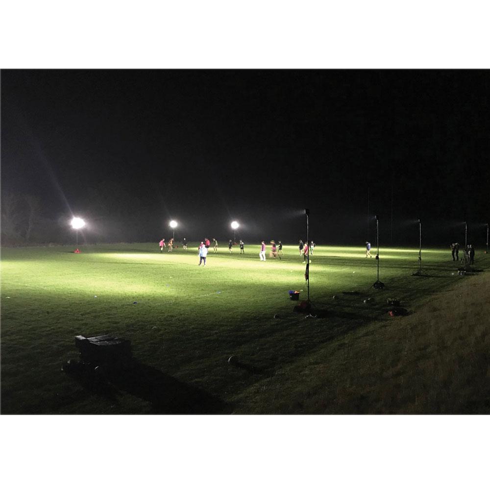 A group plays soccer at night on a grass field lit by bright 20,000-lumen Nightsearcher SportStar Portable LED Floodlights. The surroundings are dark, with equipment visible on the sidelines.