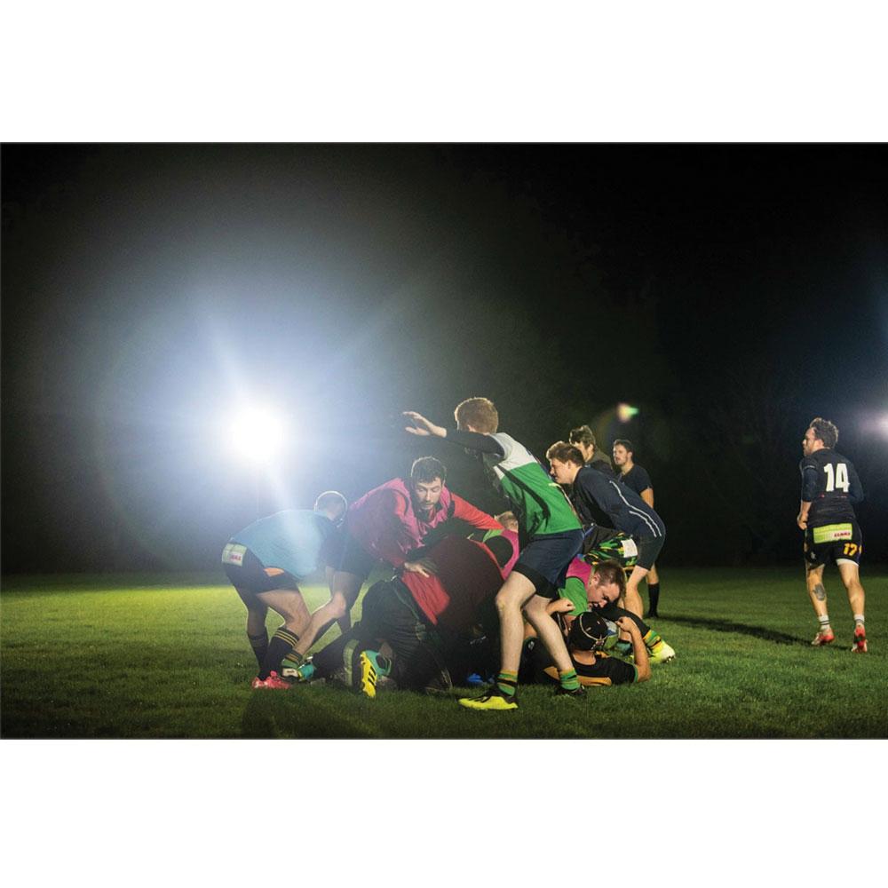 During a nighttime rugby match on grass, players compete for the ball as a Nightsearcher SportStar Portable LED Floodlight with 20,000 lumens powerfully illuminates the field in the background.