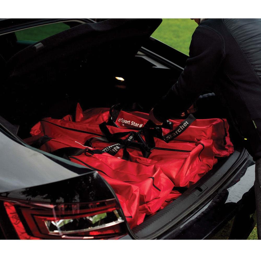 A person loads large red sports bags and a Nightsearcher SportStar Portable LED Floodlight into the open trunk of a black car, with the rear lights visible and green grass in the background.