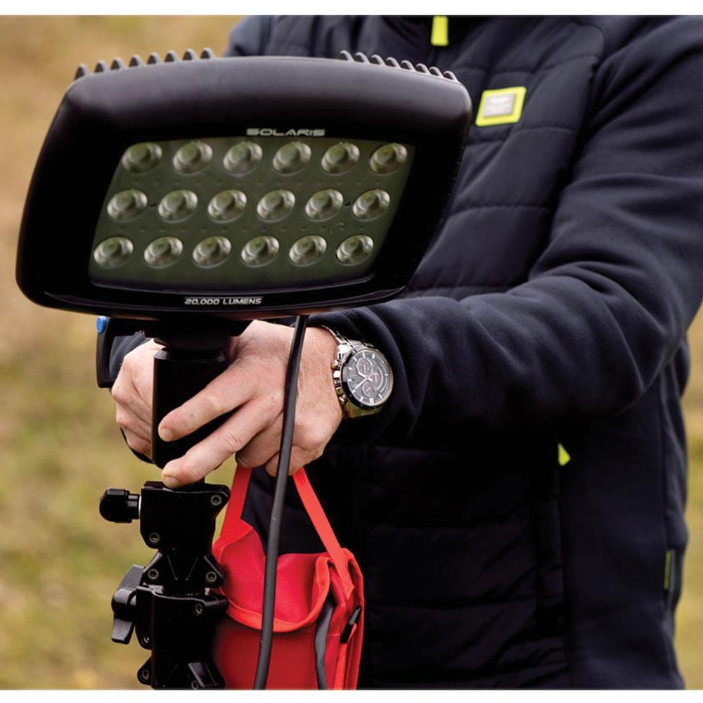 Wearing a dark jacket, a person adjusts the Nightsearcher SportStar Portable LED Floodlight outdoors. The rechargeable unit features multiple LEDs, a 20,000 LUMENS label, and a red bag hanging from its stand.