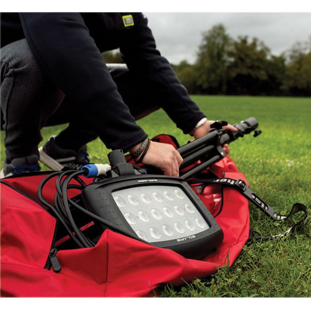 A person sets up the Nightsearcher SportStar Portable LED Floodlight (20,000 lumens) and tripod on a grassy field, with part of the light out of a red bag. Trees and cloudy skies appear in the background.