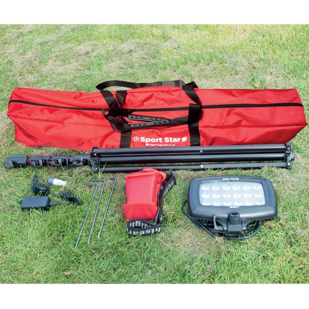 A red SportStar Portable LED Floodlight carrying case from Nightsearcher sits on grass next to a folded black tripod, 20000-lumen rechargeable floodlight, metal spikes, power adapter, and small red pouch—ideal for portable sports lighting.