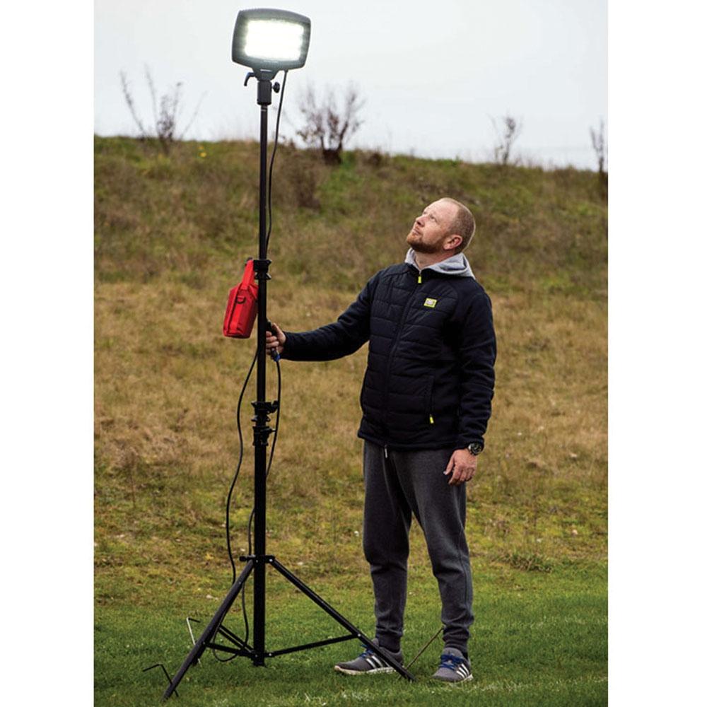 A man in a black jacket and gray sweatpants stands on grass, adjusting a Nightsearcher SportStar Portable LED Floodlight mounted on a tall tripod with a red bag attached. He looks up at the bright 20,000-lumen light under an overcast sky.