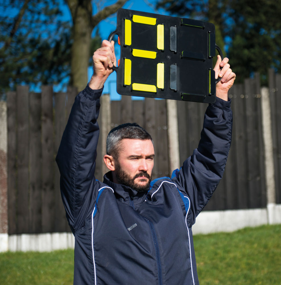 At a sports event, a man in a navy jacket holds the Precision Training Precision Substitutes Number Board displaying a bright orange 8, with a wooden fence and trees visible in the background.