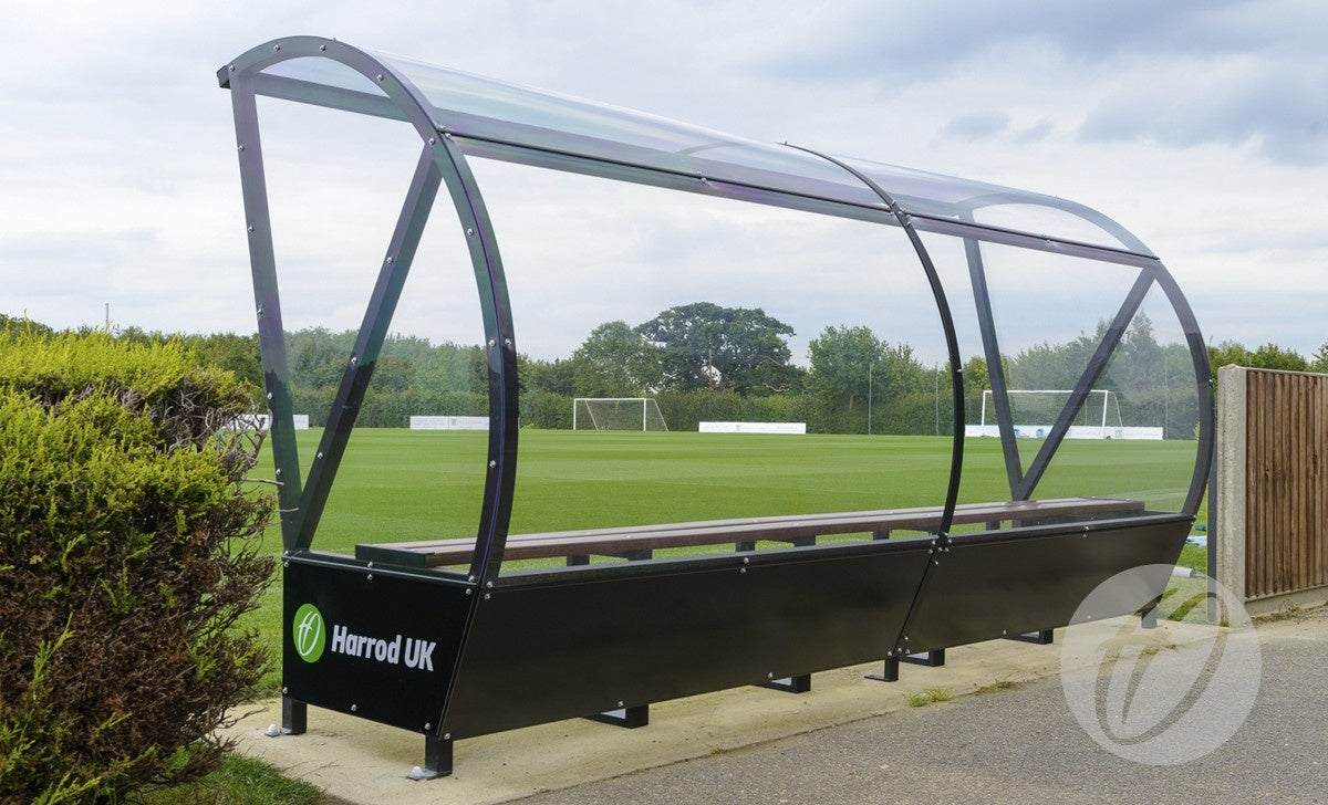 The Panoramic Team Shelter - 2M by Harrod Sport features HDPE seats and a transparent curved polycarbonate roof in a black aluminium frame, branded with Harrod Sport, positioned beside a green soccer field with trees and goalpost visible.