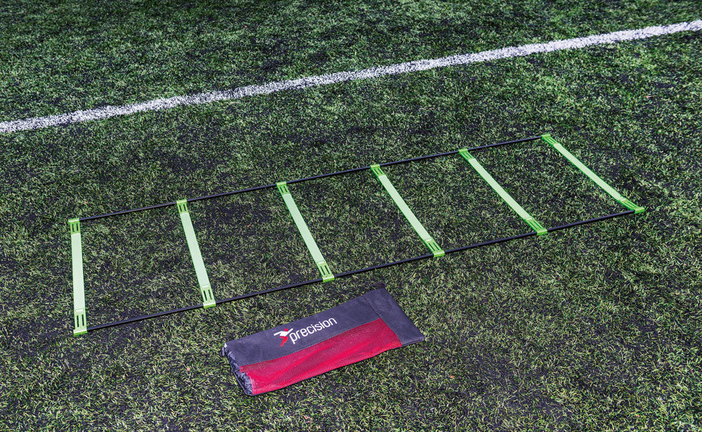 The Precision Training Pinnacle Ladder, a green and black agility ladder, with its red and black Precision bag, lies on artificial grass near a white line on the sports field.