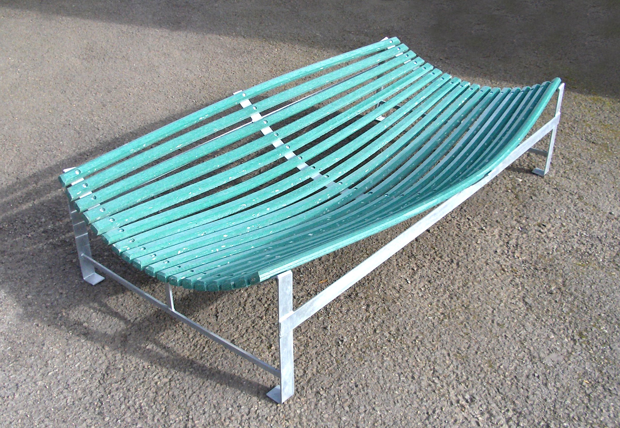 The Martin Berrill Sports Plastic Slip Catch Cradle features a curved green slatted design with a silver frame, set on a rough brown surface. Its durable structure bows inward to mimic professional fielding practice equipment.