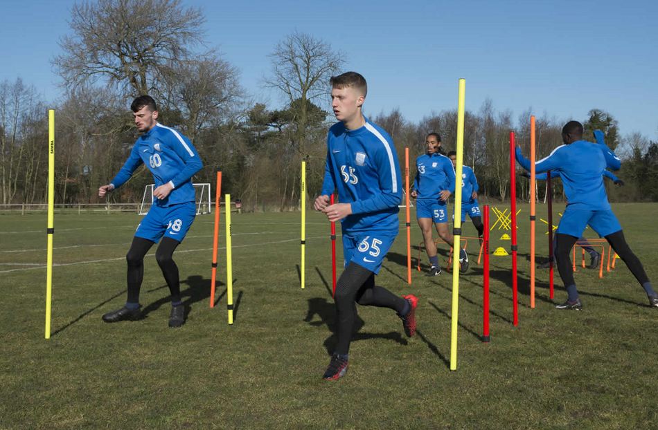 Soccer players in blue kits use Samba Sports Samba Telescopic Boundary Poles 1.7m (Set of 12) on a grassy field for speed and agility drills, with trees and clear skies in the background.
