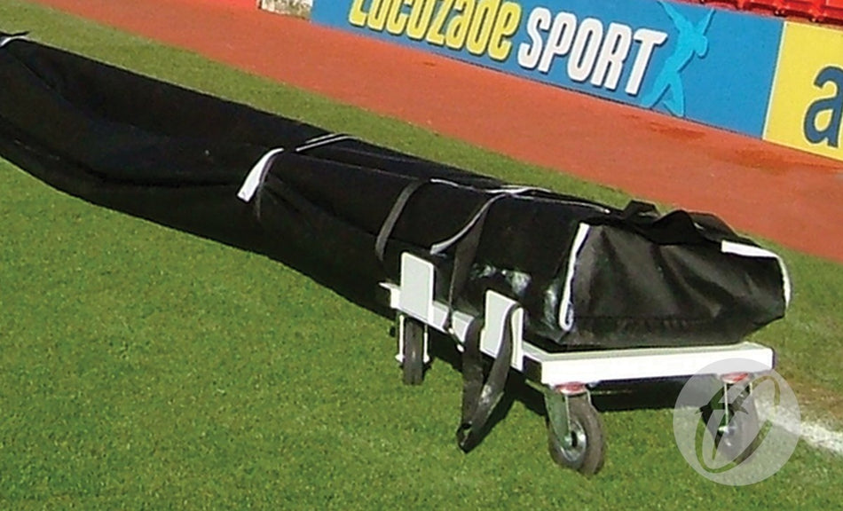 A large, black Harrod Sport equipment cover rests on a wheeled trolley on a grass field, near a stadium advertising board for Lucozade Sport. The scene also features a 3G Foldaway Euro Portagoal and a Spare 3G Stadium Foldaway Goal Bag nearby.