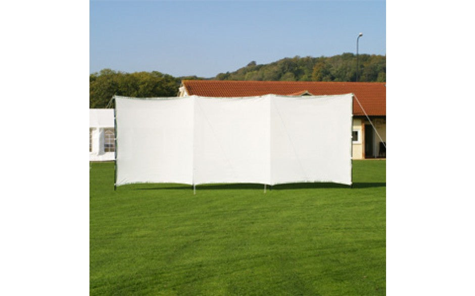 The Stuart Canvas Portable Sightscreen is positioned on a grassy lawn before a red-roofed building, with trees and hills in the background beneath a clear sky.