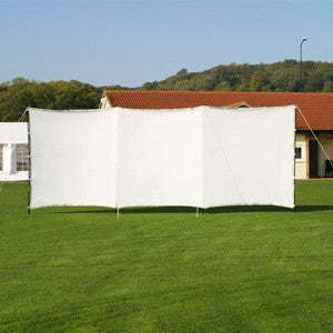 The Stuart Canvas Portable Sightscreen is set on a grassy lawn, with a building and trees in the background beneath a clear blue sky.