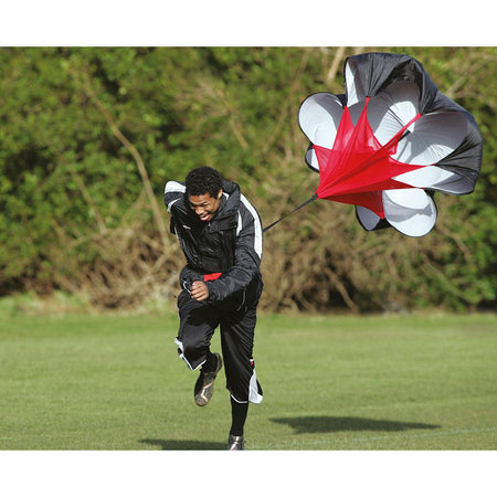 Wearing athletic clothing, a person runs on grass with the Precision Training Precision Power Speed Parachute in black, white, and red for resistance training, with trees in the background.
